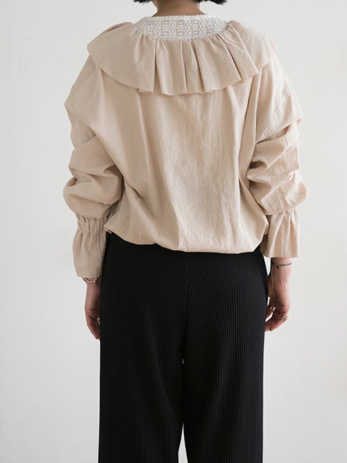 LE MELANGE（ル メランジュ）のベージュのラッフルカラーブラウスを着用した女性の後姿画像です。背中のギャザーや大きめのラッフルカラーの広がり、インナーのレースの覗き方が確認できます